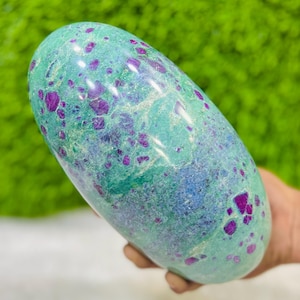 Könnte beinhalten: Ein polierter, oval geformter Edelstein mit einer türkisblauen Grundfarbe, übersät mit leuchtend magenta-farbenen Einschlüssen. Der Stein wird in einer Hand gehalten, was seine glatte, abgerundete Form zeigt. Der Hintergrund ist ein verschwommenes Grün.