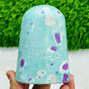 Op de afbeelding: Een turquoise stenen sculptuur met een afgeronde bovenkant en een uitlopende basis. De steen heeft onregelmatige vlekken van wit en levendige paarse insluitsels. De sculptuur wordt in een hand gehouden, met een vage groene achtergrond.