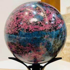 Op de afbeelding: Een gepolijste bolvormige steen met een levendige mix van diep magenta, helderblauwe en zwarte minerale insluitingen. Het gladde, ronde oppervlak vertoont natuurlijke kristalpatronen en rust op een zwarte metalen standaard met drie poten.