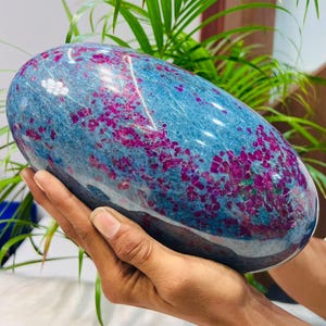 Peut inclure: Une grande pierre polie de forme ovale, lisse, présentant une couleur de base bleu profond avec des inclusions cristallines magenta et fuchsia vives et dispersées sur toute la surface, créant un aspect tacheté à fort contraste.