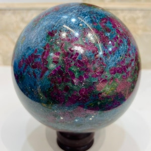 Peut inclure: Une pierre sphérique polie présentant un mélange vibrant d'inclusions minérales bleu profond et rose magenta vif sur toute sa surface. La sphère est montée sur une petite base circulaire brun foncé, affichant une texture lisse et réfléchissante.