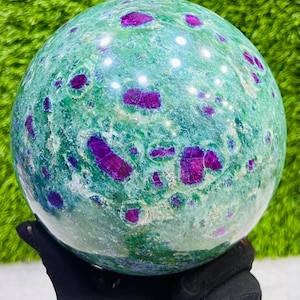 Może przedstawiać: Wypolerowana kula z kamienia szlachetnego, przeważnie zielona z żywymi fioletowymi wtrąceniami. Gładka, odbijająca światło powierzchnia uwydatnia naturalne wzory. Kula trzymana w rękawicy, podkreślając jej kształt. Unikalny przedmiot dekoracyjny.