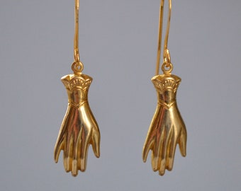 Peut inclure: Une paire de boucles d'oreilles dorées en forme de main. Les boucles d'oreilles sont suspendues à une barre dorée.