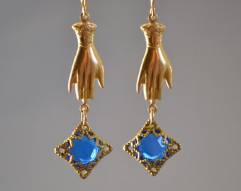 Peut inclure: Boucles d'oreilles en or avec un motif de main et une pierre précieuse bleue dans un sertissage en filigrane doré.