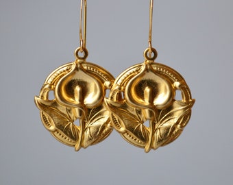 Peut inclure: Une paire de boucles d'oreilles dorées avec un motif de calla. Chaque boucle d'oreille a un cadre circulaire avec un motif détaillé de lys et de feuilles. Les boucles d'oreilles sont suspendues à de fins crochets d'oreilles dorés, pour un style classique et élégant.