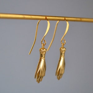 May include: A pair of gold-toned earrings featuring a hand design. The earrings are hanging from a gold-toned bar.