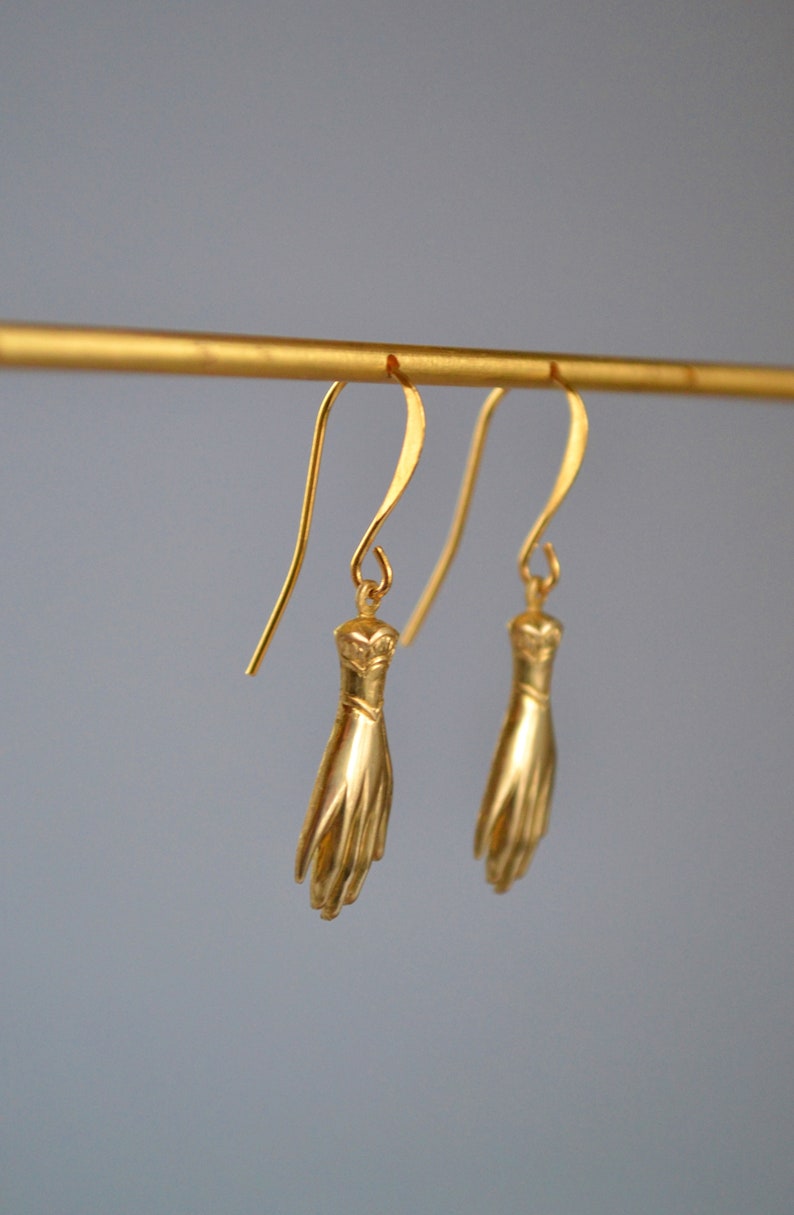 May include: A pair of gold-toned earrings featuring a hand design. The earrings are hanging from a gold-toned bar.