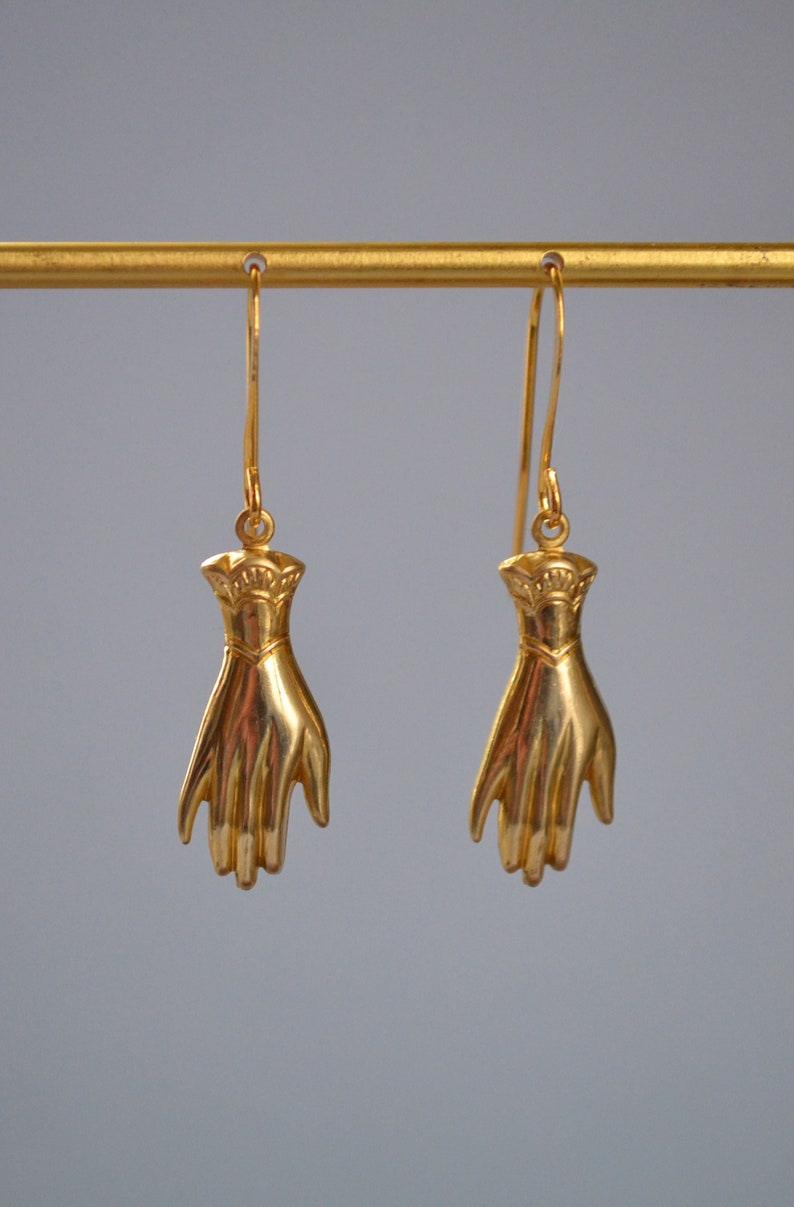 May include: A pair of gold-tone earrings featuring a hand design. The earrings are hanging from a gold-tone bar.