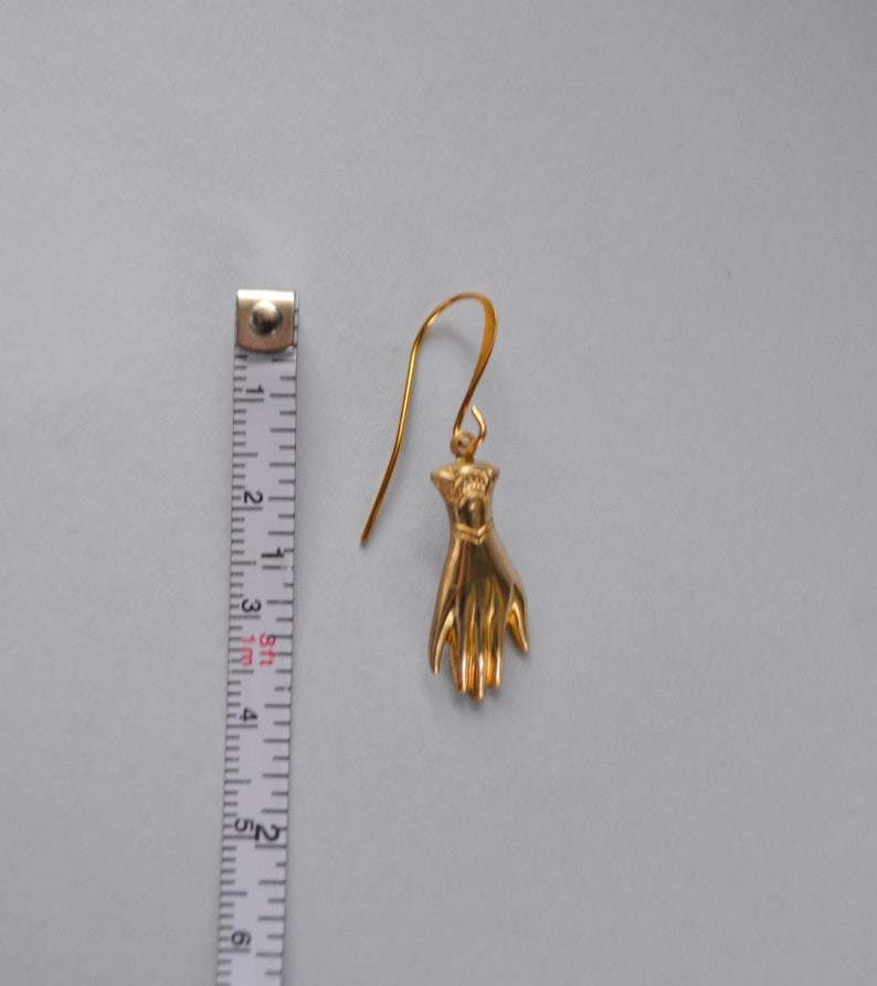 May include: Gold-toned hand-shaped dangle earring with a hook closure. The earring is approximately 2 inches long.