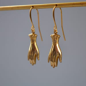 May include: A pair of gold-tone earrings featuring a hand design. The earrings are hanging from a gold-tone bar.