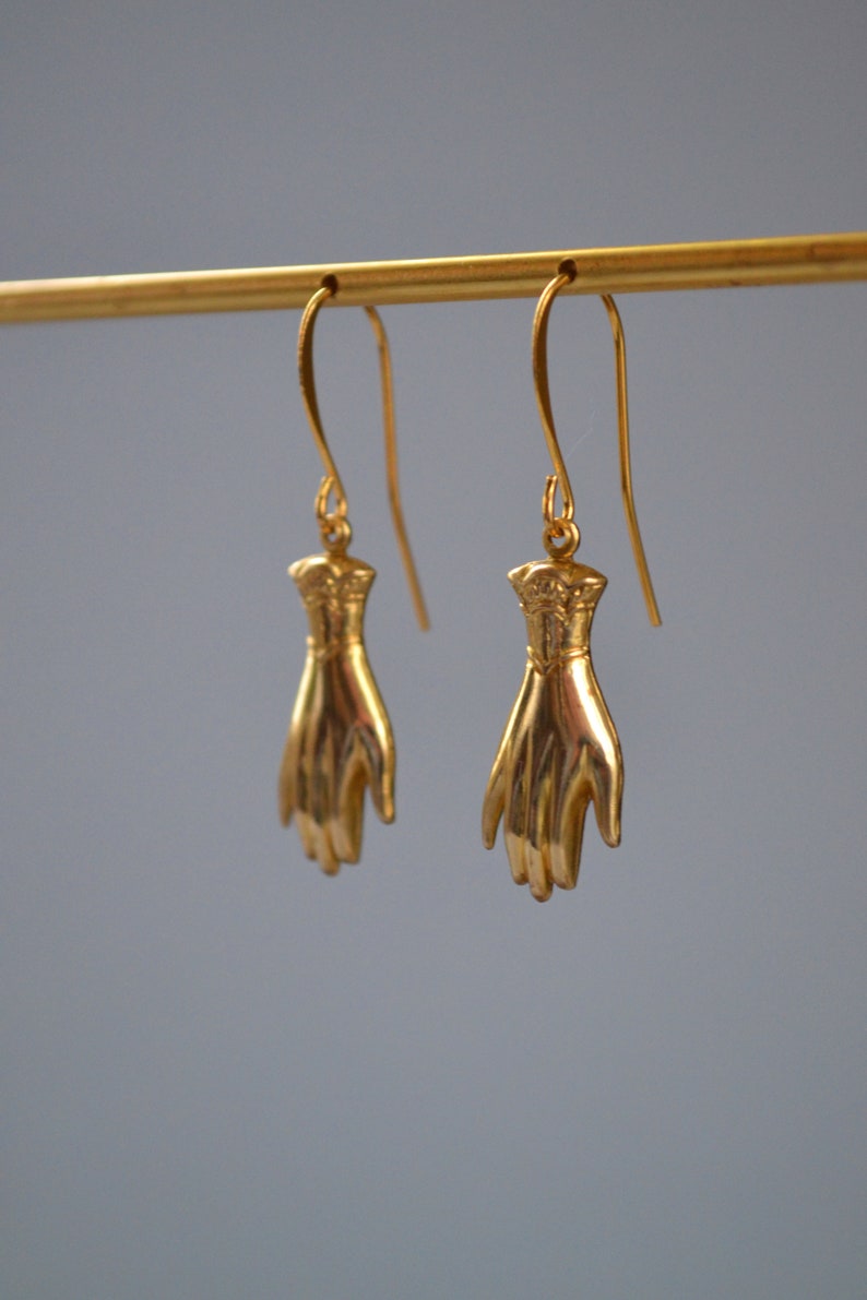 May include: A pair of gold-tone earrings featuring a hand design. The earrings are hanging from a gold-tone bar.