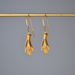 May include: A pair of gold-toned earrings featuring a hand design. The earrings are hanging from a gold-toned bar.