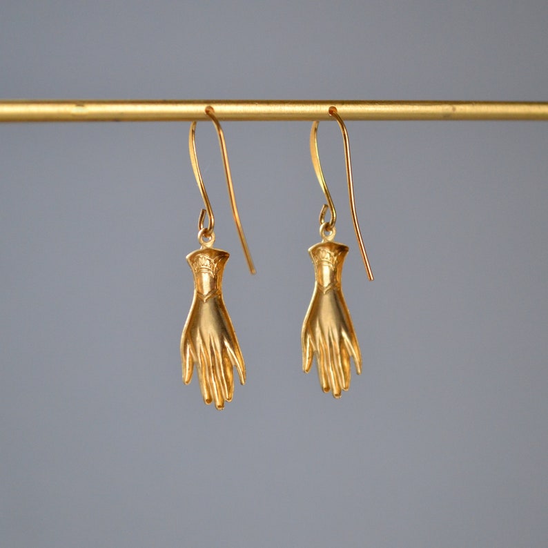 May include: A pair of gold-toned earrings featuring a hand design. The earrings are hanging from a gold-toned bar.