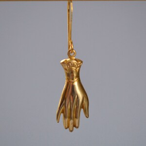 May include: Gold-toned hand-shaped dangle earrings. The hand is detailed with a textured surface and a raised design on the wrist.