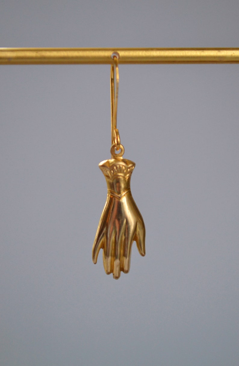 May include: Gold-toned hand-shaped dangle earrings. The hand is detailed with a textured surface and a raised design on the wrist.