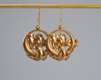 Peut inclure: Une paire de boucles d'oreilles dorées. Chaque boucle d'oreille présente un motif floral dans un cercle. Les boucles d'oreilles sont suspendues à un fin fil d'or et sont présentées sur un fond neutre.