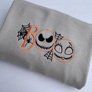 May include: A light gray sweatshirt with an embroidered Halloween design. The design features the word "BOO" in orange, with a spider web and two cartoon faces. One face has a sinister expression, and the other has a smiling expression.