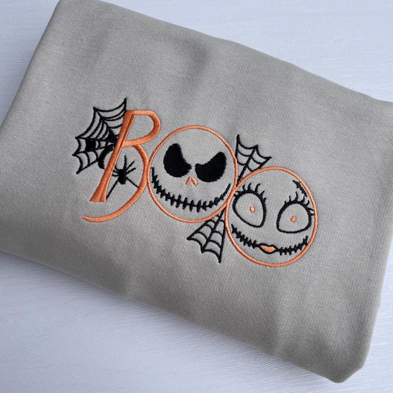 May include: A light gray sweatshirt with an embroidered Halloween design. The design features the word "BOO" in orange, with a spider web and two cartoon faces. One face has a sinister expression, and the other has a smiling expression.