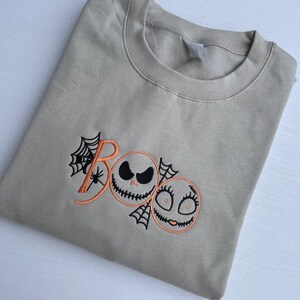 May include: A tan-colored sweatshirt with the word "Boo" embroidered in orange, featuring Jack Skellington and Sally faces. Black spiderwebs and a spider are also embroidered on the sweatshirt. The sweatshirt is folded.