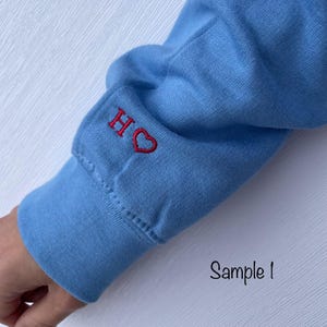 Op de afbeelding: Een lichtblauw shirt met lange mouwen met een rood geborduurd hart en de letter "H" op de manchet.