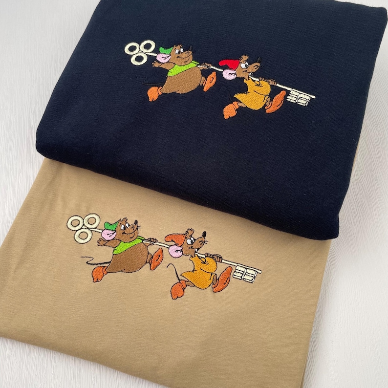 May include: Two tan t-shirts with embroidered images of three mice carrying a large key. The mice are wearing different colored hats. One shirt is navy blue and the other is tan.