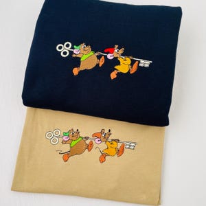 May include: Two embroidered mice holding a key on a tan t-shirt. The mice are wearing green and yellow shirts. The t-shirt is on top of a black t-shirt.