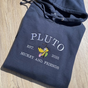 May include: A navy blue hoodie featuring the embroidered word "PLUTO" in white. Below is an embroidered image of Pluto, the Disney character, with the text "EST. MICKEY AND FRIENDS 2025". The hoodie has a drawstring hood.