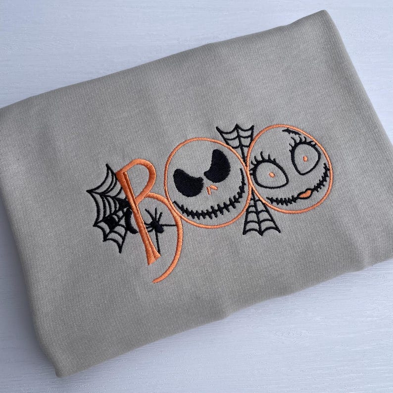 May include: A light gray sweatshirt with an embroidered Halloween design. The word "BOO" is spelled out in orange thread, with the "O"s replaced by Jack Skellington and Sally faces. Black spiderwebs and a spider are also embroidered.