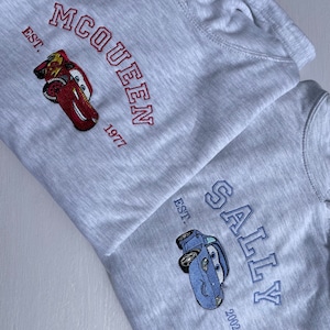 May include: Two grey hooded sweatshirts with embroidered logos. The top sweatshirt has "McQueen EST. 1977" in red with a red cartoon car. The bottom sweatshirt has "Sally EST. 2002" in blue with a blue cartoon car.