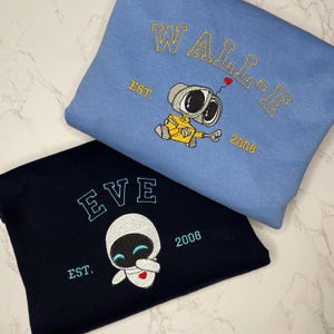 May include: Two sweatshirts, one light blue and one navy blue, with embroidered designs. The light blue sweatshirt features "WALL-E" and a robot design, with "EST. 2008." The navy blue sweatshirt features "EVE" and a robot design, with "EST. 2008."