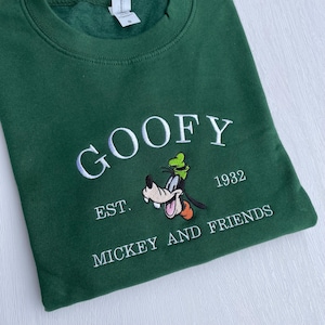 May include: A dark green sweatshirt featuring the embroidered word "GOOFY" in white. Below the word "GOOFY" is an embroidered image of Goofy, the Disney character, and the text "EST. 1932 MICKEY AND FRIENDS".