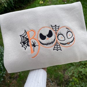 May include: A light gray sweatshirt with an embroidered Halloween design. The design features the word "BOO" in orange, with a spider web on the "B". Two cartoon faces, one with a sinister grin and the other with eyelashes, are also embroidered.