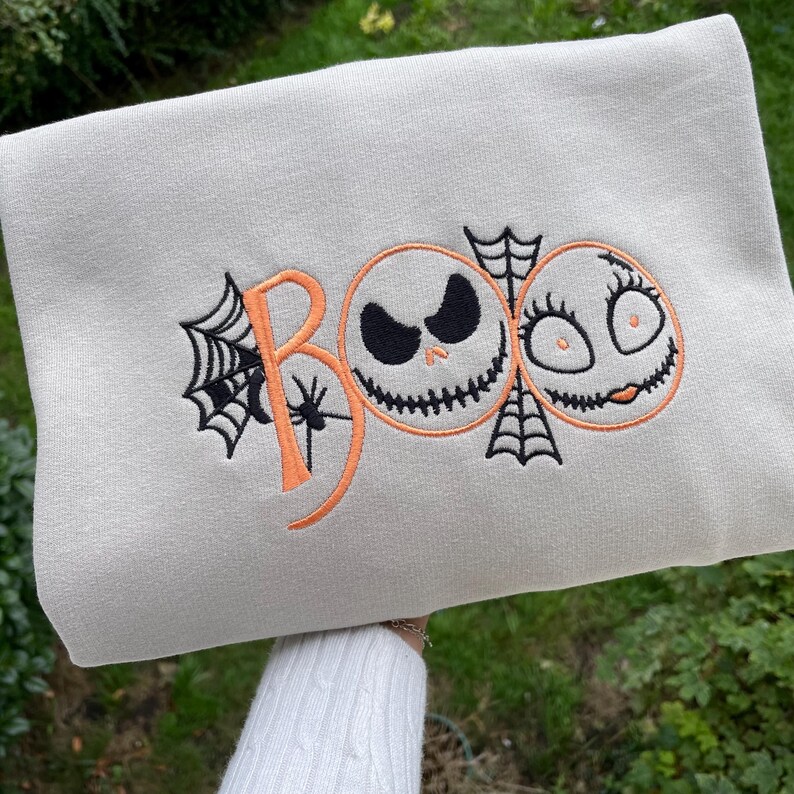 May include: A light gray sweatshirt with an embroidered Halloween design. The design features the word "BOO" in orange, with a spider web on the "B". Two cartoon faces, one with a sinister grin and the other with eyelashes, are also embroidered.
