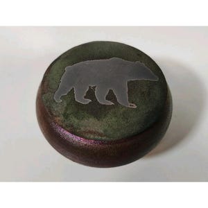 May include: A round wooden box with a dark green top and a grey bear silhouette. The box has a dark brown, almost purple, base. The bear is walking to the right. The box is on a white surface.