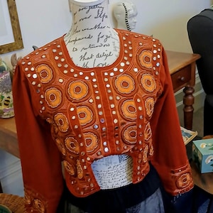 Puede incluir: Chaqueta color óxido con intrincados bordados florales naranjas y pequeños detalles de espejo. La chaqueta se exhibe en un maniquí, mostrando su diseño detallado y mangas largas. Una prenda de vestir única.