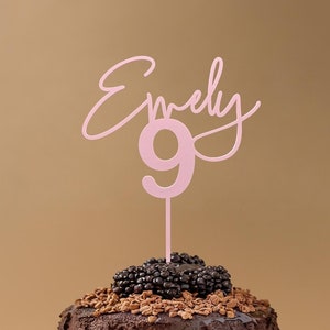 Könnte beinhalten: Ein Schokoladenkuchen mit Brombeeren und einem rosa Tortenaufsatz mit der Aufschrift "Emily 9". Der Kuchen steht auf einem schwarzen Teller vor einem beigen Hintergrund. Ein Dessert für eine Geburtstagsfeier.