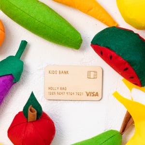 May include: A wooden toy credit card with the text "KIDS BANK" and "VISA" printed on it. The card has a name and number printed on it. The card is surrounded by felt fruit toys, including a green banana, a yellow banana, a red apple, a purple eggplant, a yellow lemon, a red watermelon, and a yellow pear.