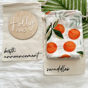 May include: Two white muslin bags with black text. One bag says "birth announcement" and the other says "swaddles". A wooden circle with the text "Holly Rao" is in the top bag. The bottom bag has a white muslin swaddle with a pattern of orange fruit and green leaves.