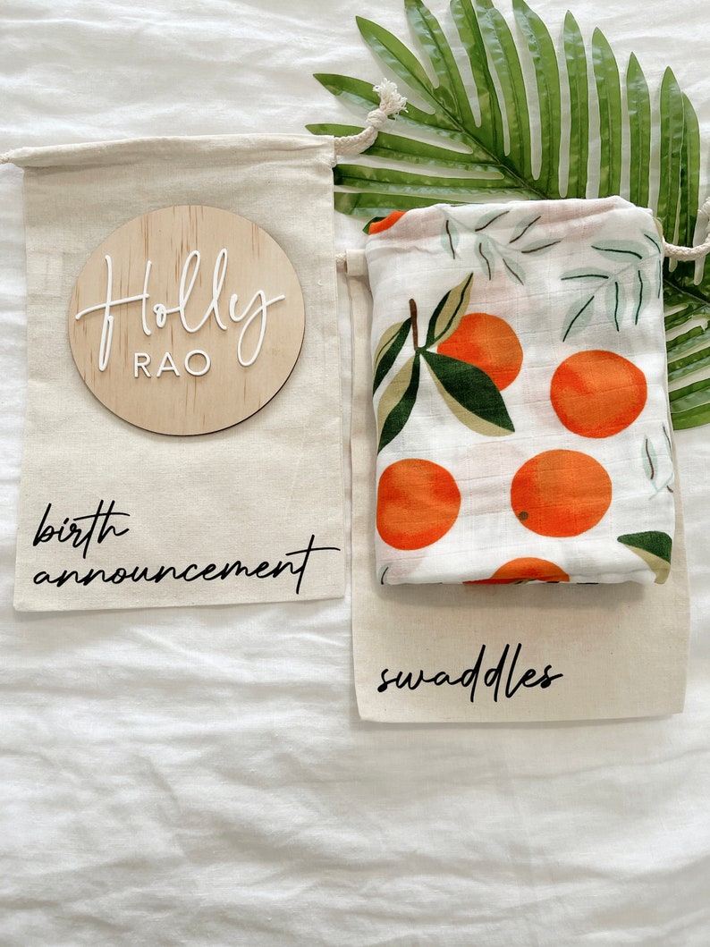 May include: Two white muslin bags with black text. One bag says "birth announcement" and the other says "swaddles". A wooden circle with the text "Holly Rao" is in the top bag. The bottom bag has a white muslin swaddle with a pattern of orange fruit and green leaves.