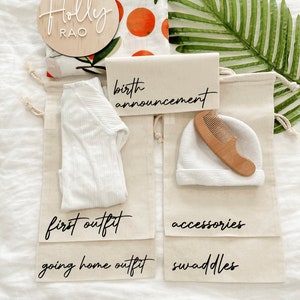 May include: A set of four white canvas bags with black lettering. The bags are labeled "birth announcement", "first outfit", "going home outfit", and "swaddles". There is a wooden sign with the name "Holly Rao" and a white onesie, a white hat, and a wooden comb inside the bags.