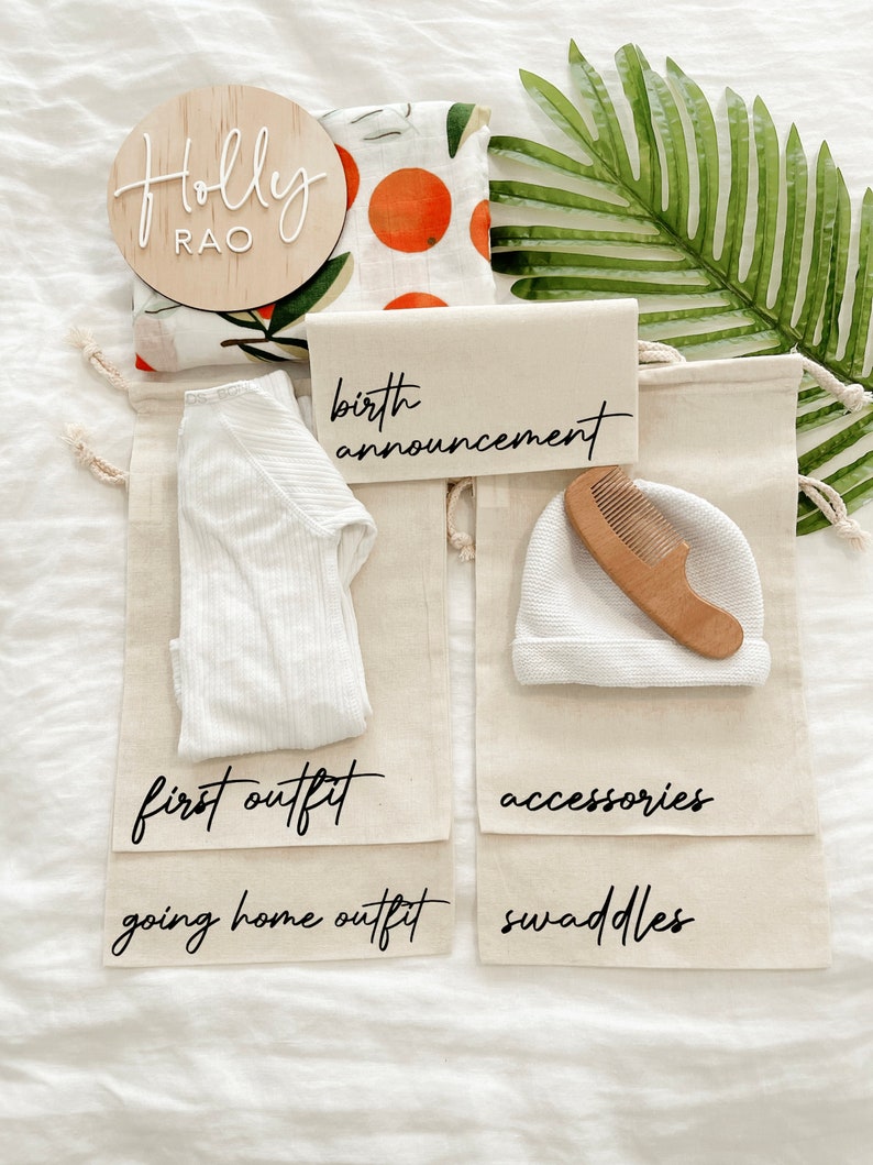 May include: A set of four white canvas bags with black lettering. The bags are labeled "birth announcement", "first outfit", "going home outfit", and "swaddles". There is a wooden sign with the name "Holly Rao" and a white onesie, a white hat, and a wooden comb inside the bags.