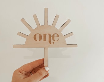 May include: Wooden sunburst cake topper with the word "one" in the center. The topper is light brown and has a simple design.