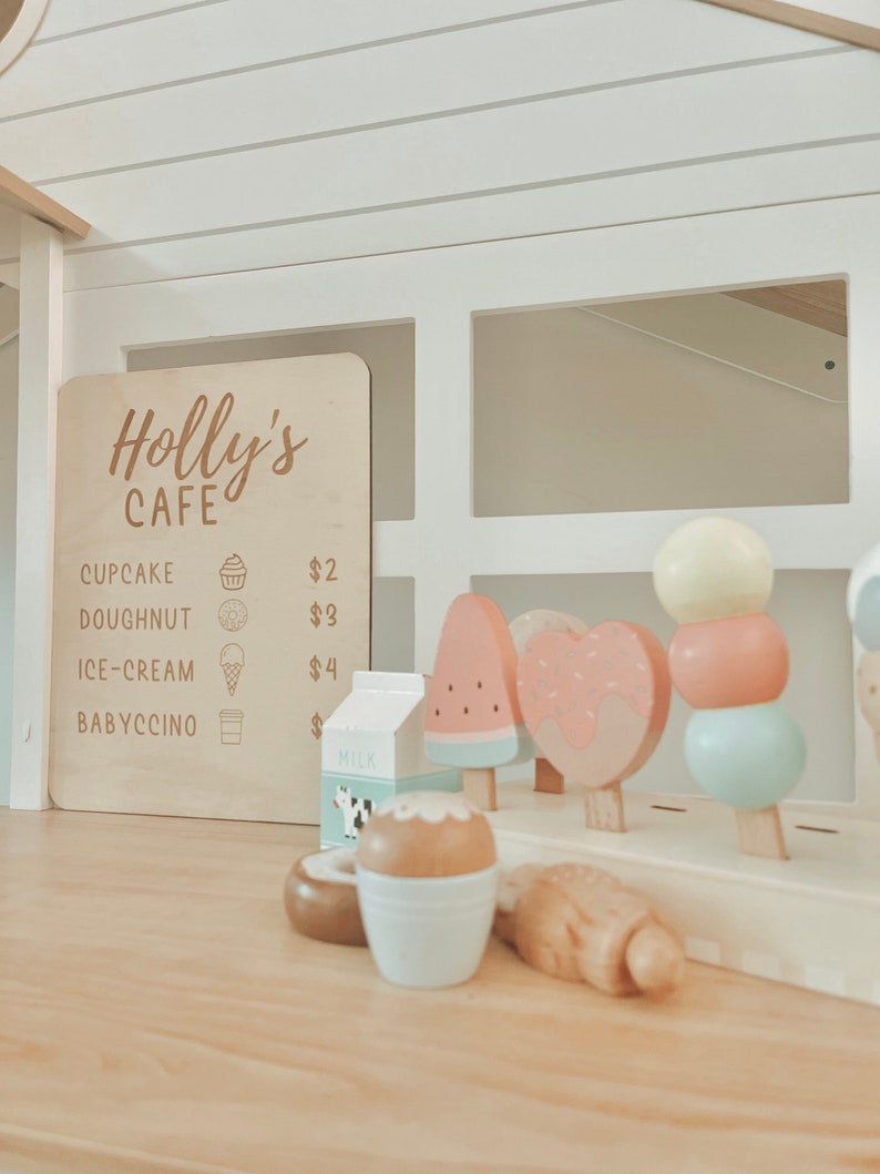 May include: A wooden play cafe sign with the text "Holly's Cafe" and a menu listing prices for cupcake, doughnut, ice cream, and babyccino.  The sign is displayed on a white wooden play counter with a variety of wooden play food items, including a watermelon popsicle, a heart-shaped donut, a popsicle with three round balls, and a carton of milk.