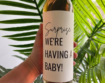 May include: A clear wine bottle with a white label that reads "Surprise WE'RE HAVING A BABY!" in black script. The bottle is held in front of a green leafy background. The wine inside appears to be a light rose color.
