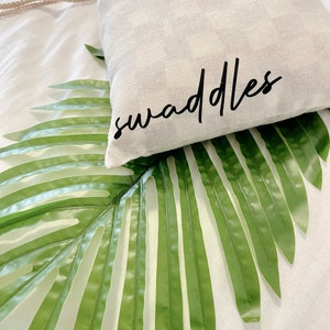 May include: A white fabric bag with the word "swaddles" printed in black lettering. The bag is lying on a green palm leaf.