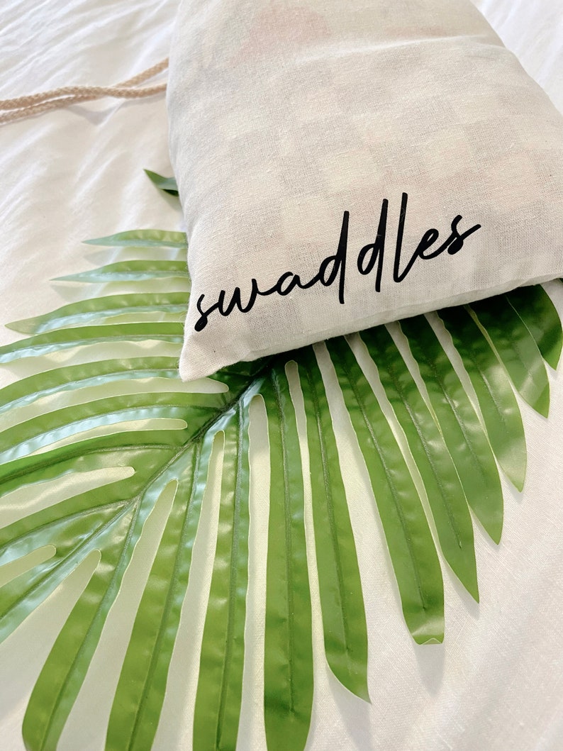 May include: A white fabric bag with the word "swaddles" printed in black lettering. The bag is lying on a green palm leaf.