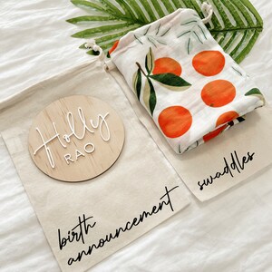 May include: A white muslin swaddle with an orange citrus print, a wooden round sign with the name "Holly Rao" and the text "birth announcement" and "swaddles" on a white fabric background.