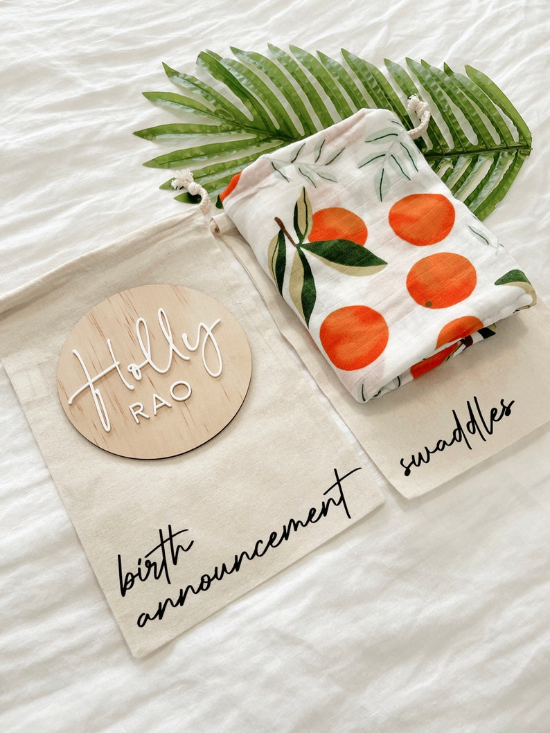 May include: A white muslin swaddle with an orange citrus print, a wooden round sign with the name "Holly Rao" and the text "birth announcement" and "swaddles" on a white fabric background.
