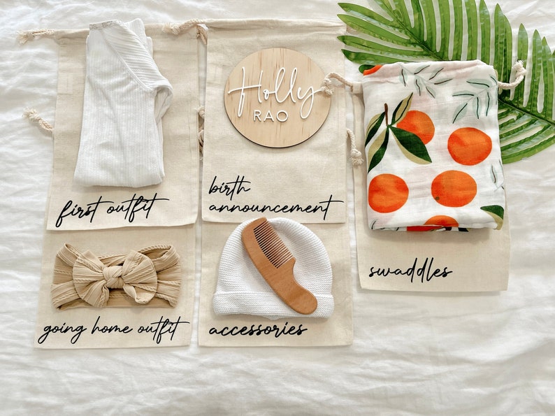 May include: Six white canvas bags with black text labels. The bags contain baby items, including a white onesie, a headband, a white hat, a wooden comb, and a swaddle with an orange citrus print. The bags are labeled "first outfit", "birth announcement", "going home outfit", "accessories", "swaddles", and a wooden circle with the text "Holly Rao".