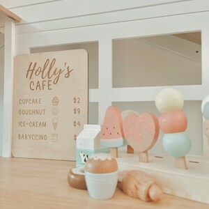 May include: A wooden play cafe sign with the text "Holly's Cafe" and a menu listing prices for cupcake, doughnut, ice cream, and babyccino. The sign is displayed on a white wooden counter with a variety of wooden play food items, including a watermelon popsicle, a donut, and a popsicle.